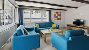 Ett vardagsrum med blå soffor, ett soffbord i trä, en tv på ett träställ och ett fönster med gardiner.