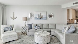 Un salon avec un canapé d’angle gris, une table basse ronde et un tableau représentant des voiliers.