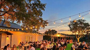 An outdoor dining area with string lights, tables set for guests, and a building in the background. - Gated 10-Acre Compound | Sleeps 28 | Game Room (Spicewood)