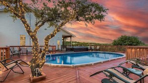A wooden deck with a swimming pool, lounge chairs, and a tree wrapped with lights. - Gated 10-Acre Compound | Sleeps 28 | Game Room (Spicewood)