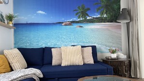 Een woonkamer met een blauwe bank, een glazen salontafel en een muurschildering van een tropisch strand.