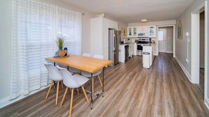 Een moderne keuken met witte kastjes, een houten eettafel en een groot raam met dunne gordijnen.