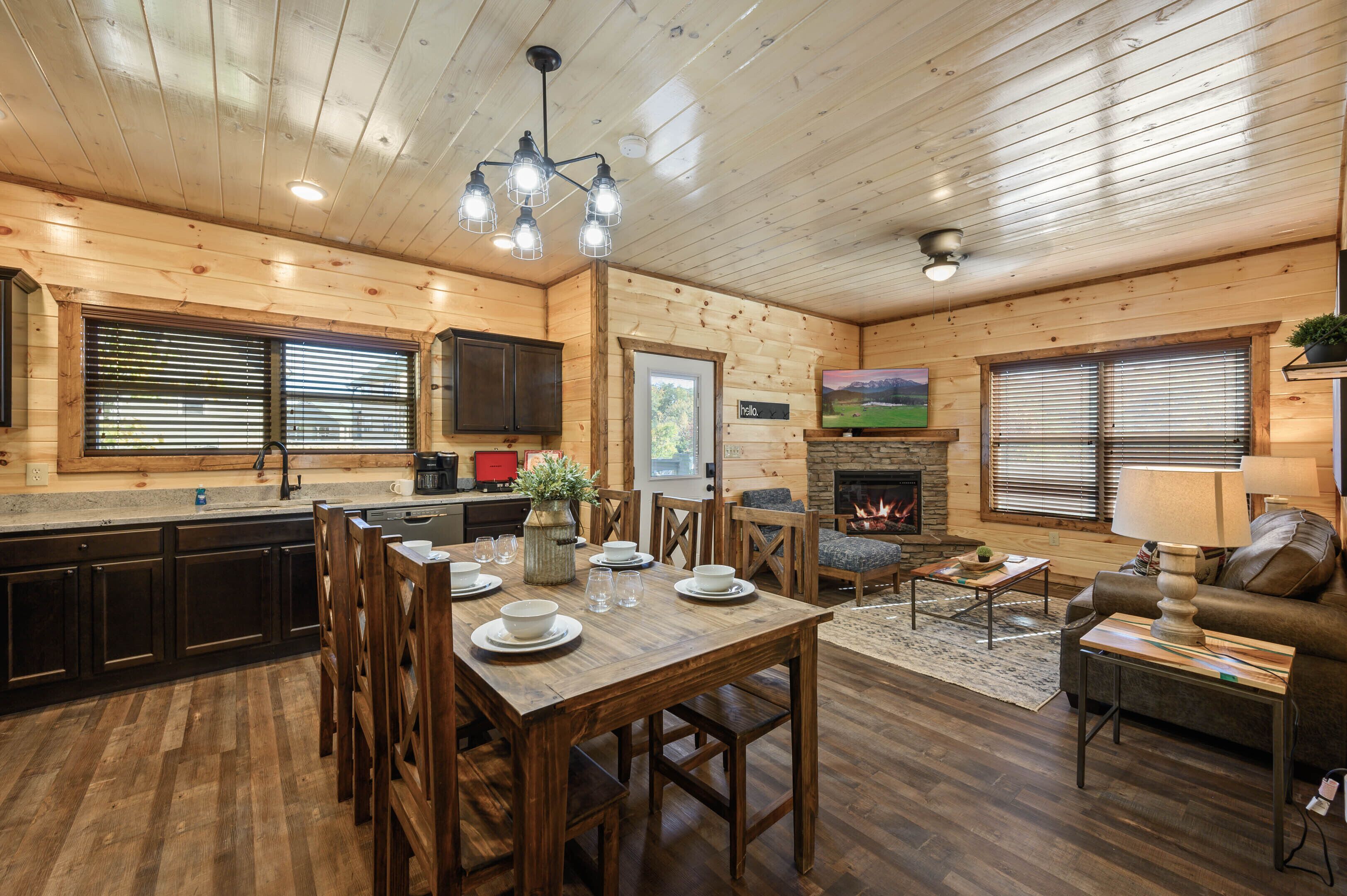 A wooden cabin interior with a dining area, kitchen, living room, and fireplace.