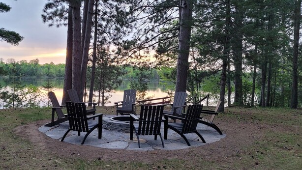 A circular outdoor seating area with Adirondack chairs and a fire pit, surrounded by tall trees and a lake view.