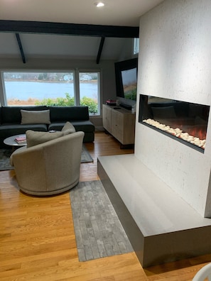 Uma sala de estar moderna com lareira, um sofá, uma mesa de centro e vista para um lago através de grandes janelas.