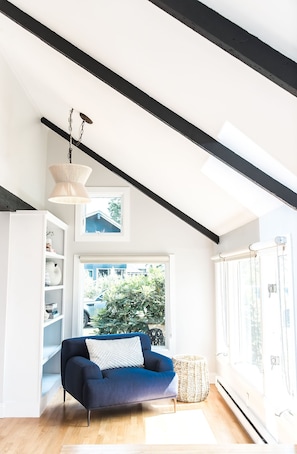Uma sala de estar moderna com um sofá azul, uma estante branca e uma janela grande com vista para o verde e um edifício.