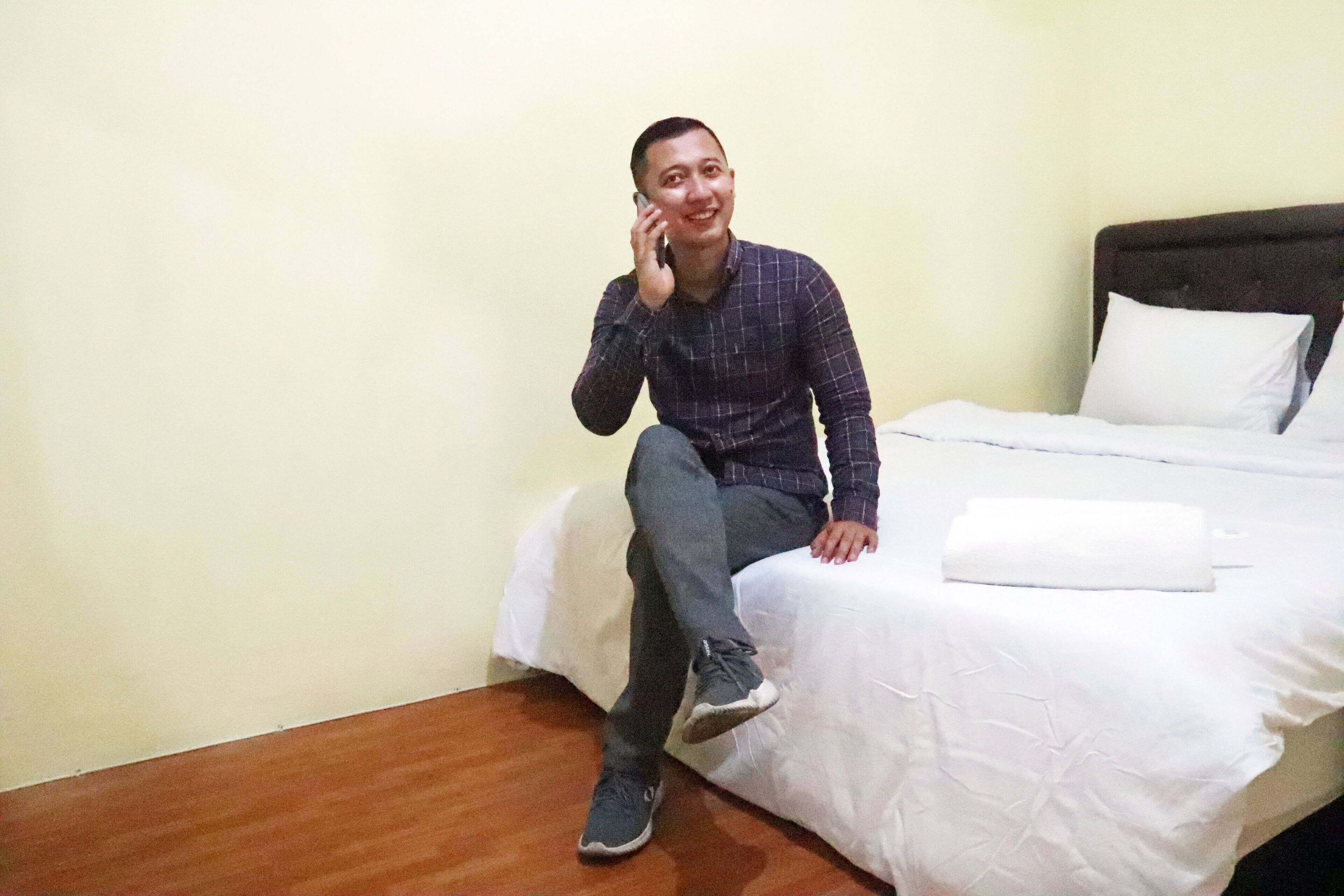 Un uomo seduto su un letto in una camera d'albergo con una parete spoglia e un pavimento in legno.