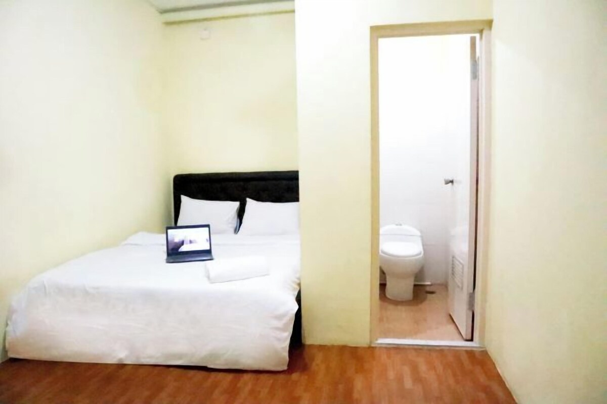 Una habitación pequeña con una cama individual, suelo de madera, colcha blanca y mesita de noche con un libro.