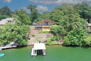 A two-story house with a balcony, surrounded by trees and a dock extending into a green body of water.