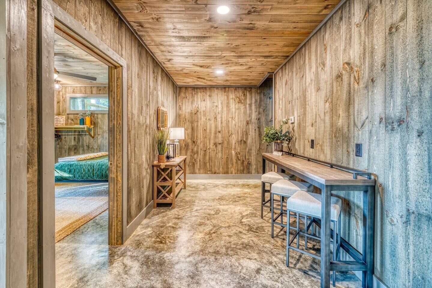 A rustic interior with wooden walls and ceiling, a small table with a lamp, and a bar area with stools.