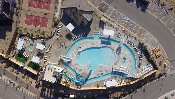 Vista aerea di un complesso piscine con scivolo acquatico, lettini prendisole e campo da tennis.