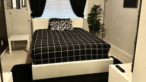 A bedroom with a bed, a bedside table, a mirror, a plant, and a wall with a sign.