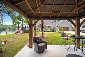 A covered patio area with a wicker chair and a table, overlooking a lake and residential buildings.