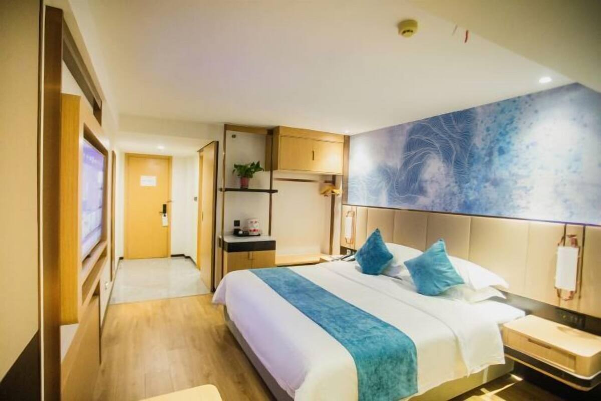 A hotel room with a bed, bedside tables, a nightstand, a desk, a chair, a shelf with a plant, and a wall-mounted artwork.