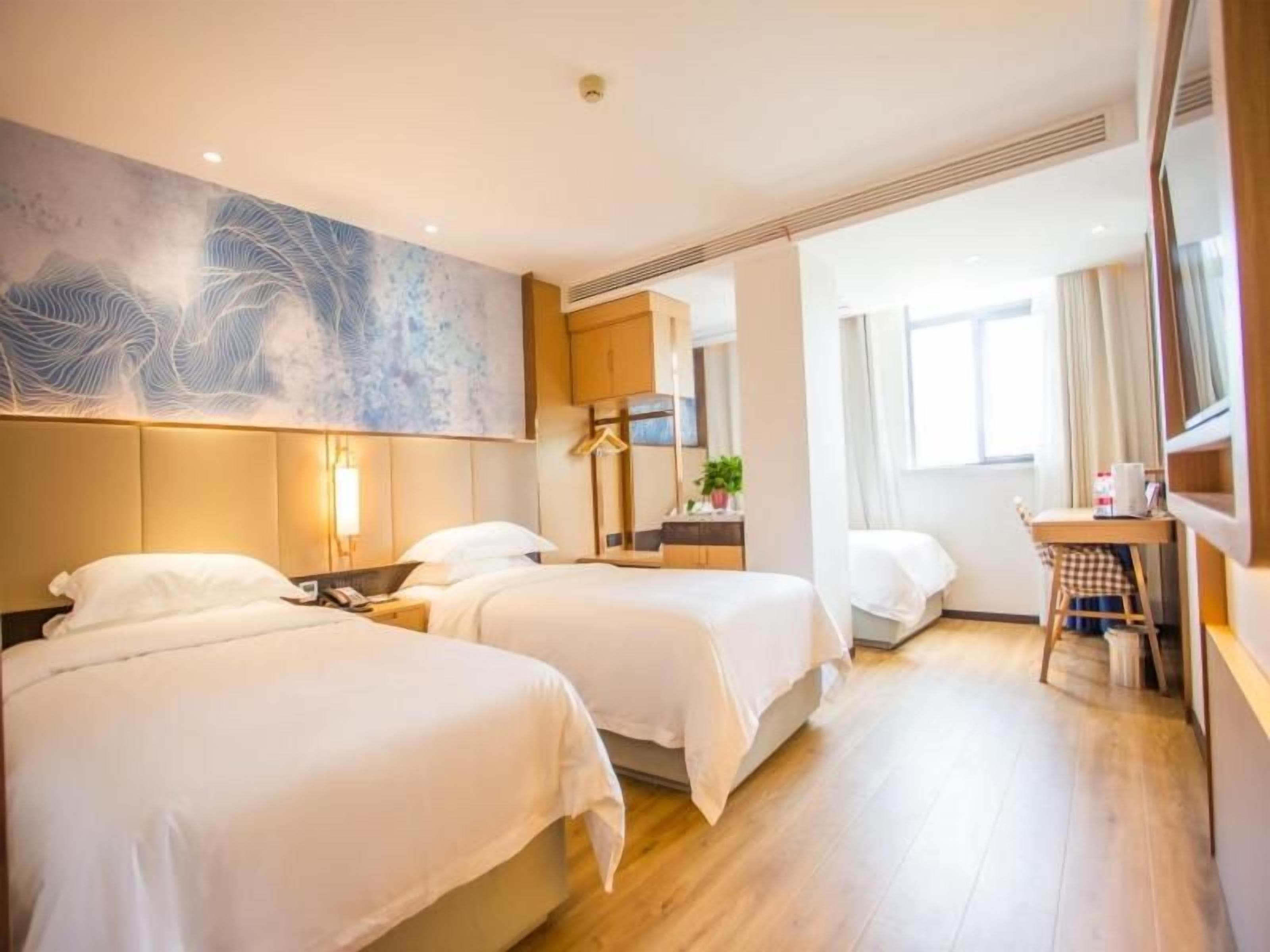 Et hotelværelse med to senge, et skrivebord, en stol, et stort vindue og et kunstværk på væggen.