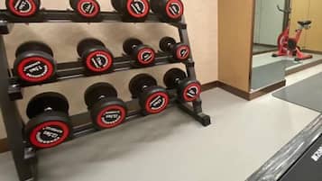 Un estante con mancuernas y sus indicaciones de peso, una bicicleta estática roja y un espejo que refleja parte de la habitación.