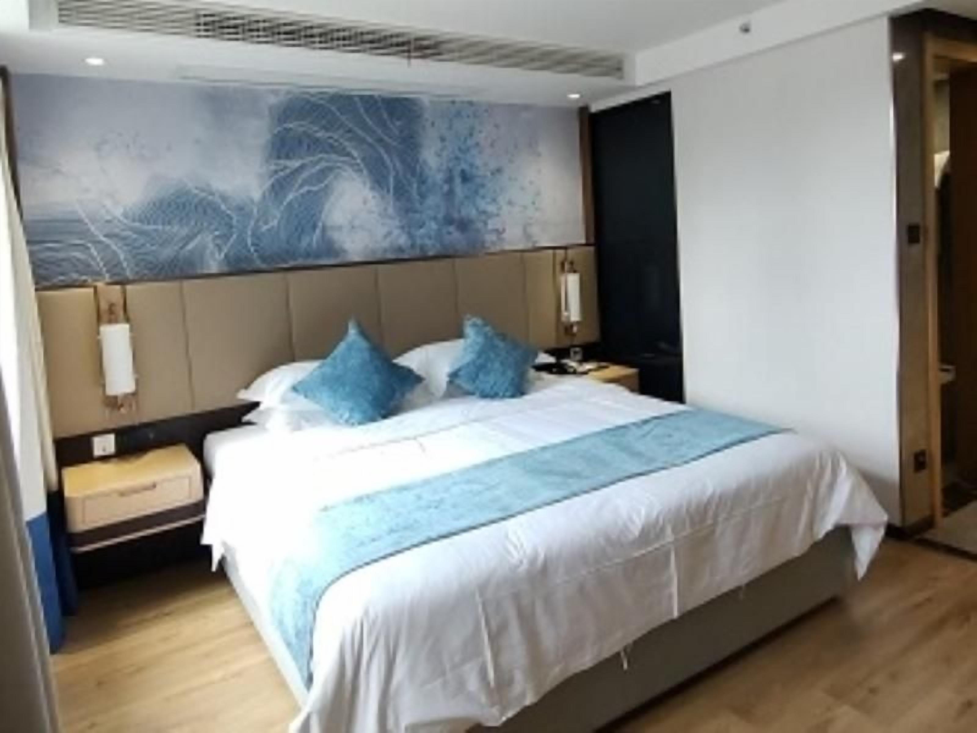 Een hotelkamer met een groot bed, een hoofdeinde met een lichtgekleurd paneel, een nachtkastje met een lamp en een aan de muur bevestigd kunstwerk.