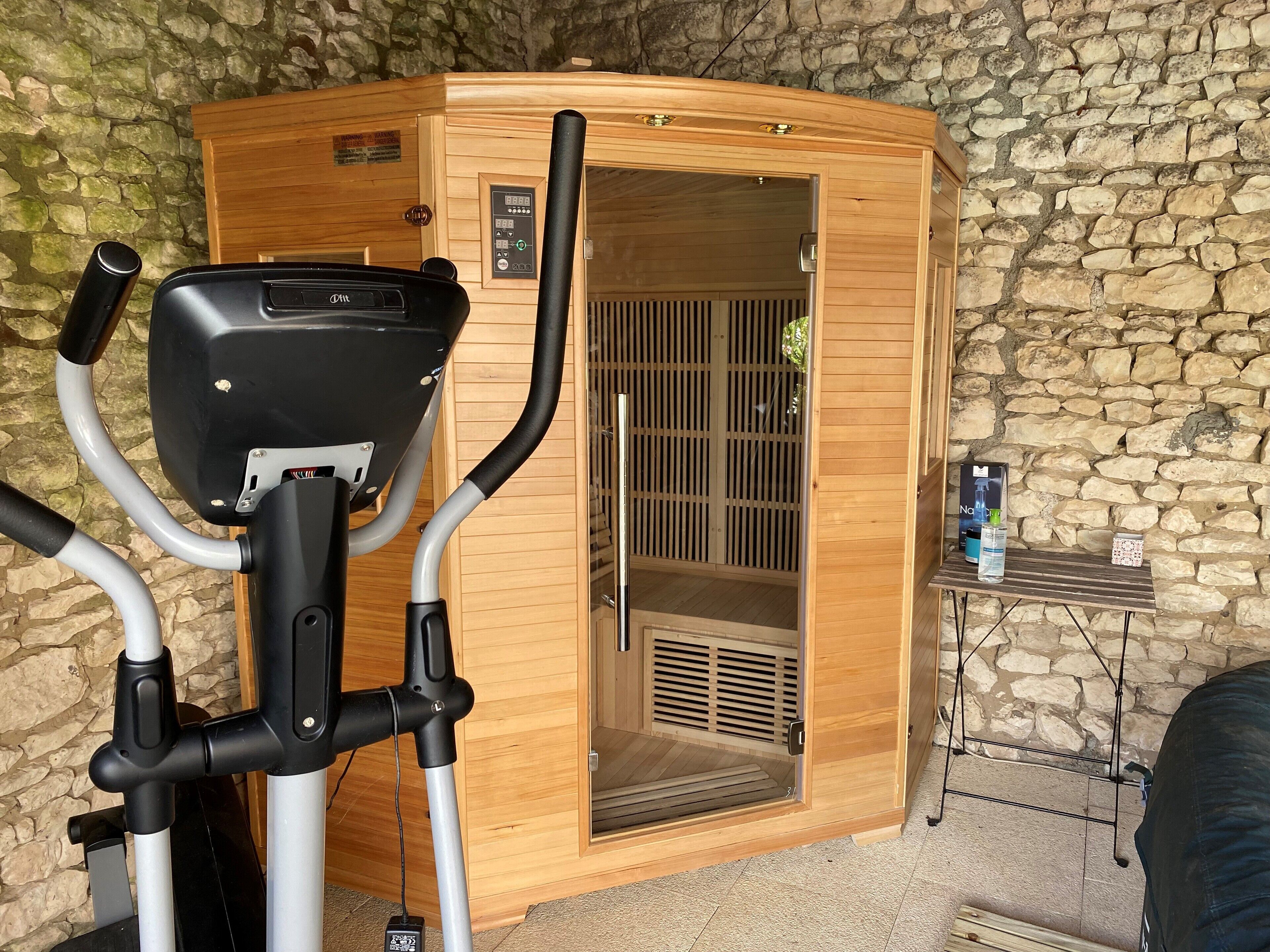 Una sauna de madera con un panel de control, un banco de madera en el interior y una pequeña mesa con una botella de agua y una caja a un lado.