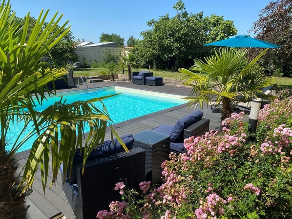 A swimming pool with a wooden deck, surrounded by lush greenery and pink flowers. - Gite Le Travezay avec piscine, sauna et Jacuzzi (Courcoué)