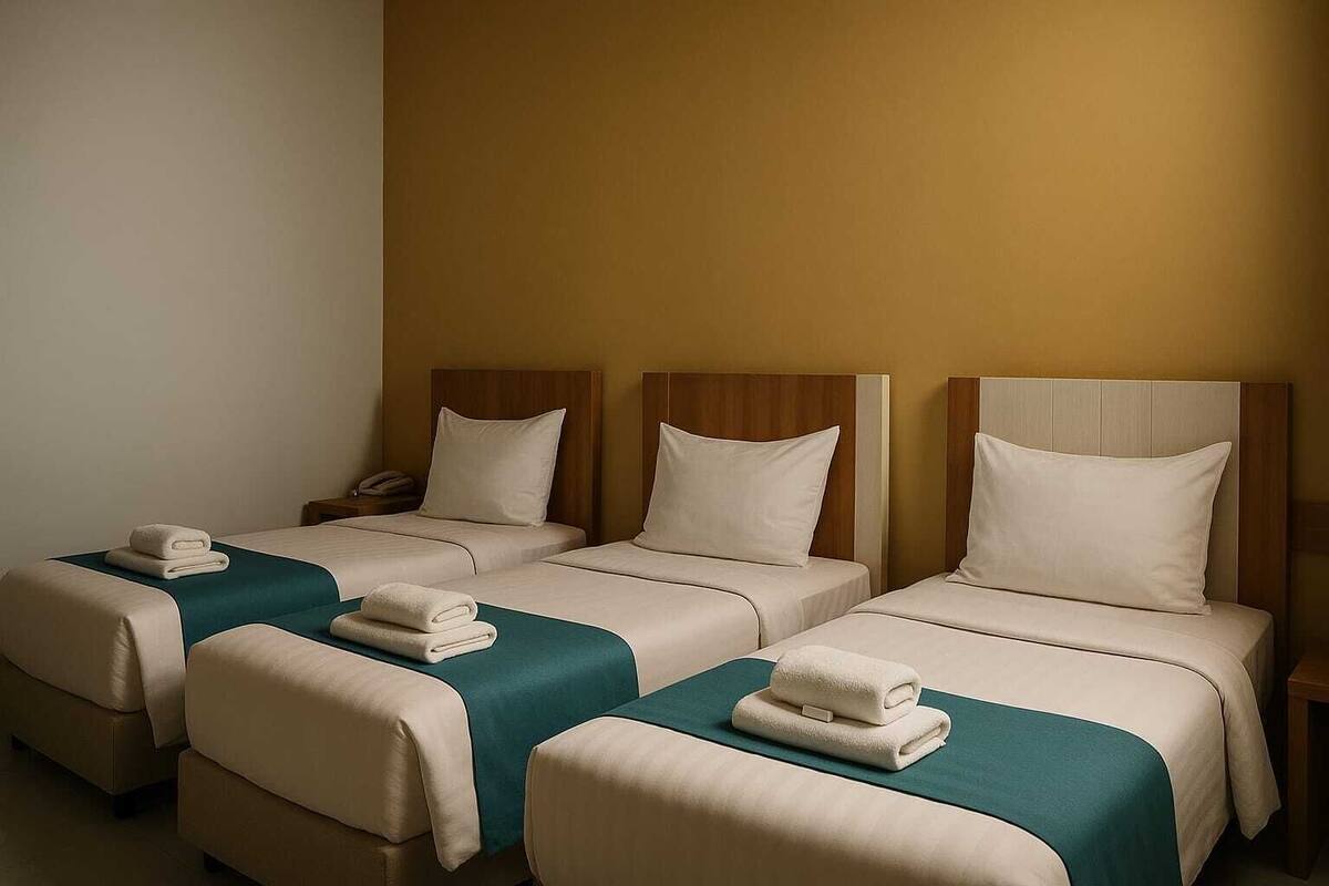 A hotel room with three single beds, each with a wooden headboard, a teal bedspread, and a white pillow. There are folded towels on each bed.
