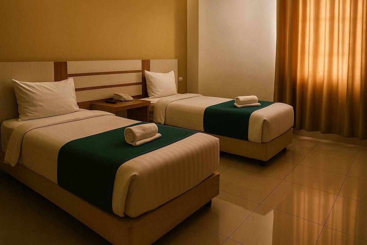 A hotel room with two beds, each with a green blanket and a rolled towel. There is a wooden headboard, a nightstand with a phone, and a window with curtains.