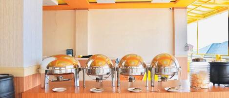 Una mesa de buffet con cuatro recipientes grandes de comida, un dispensador de agua y platos apilados.