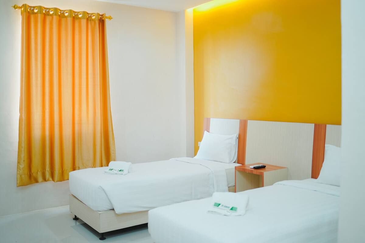 A hotel room with two beds, a curtain, a bedside table with a remote control, and a bright wall.