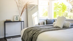 Ein Schlafzimmer mit einem Bett, einem Nachttisch mit Büchern und einer Vase, einem Spiegel und einem Fenster mit Blick auf Bäume.