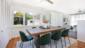 Ein Essbereich mit einem Holztisch, grünen Stühlen, einem Deckenventilator und großen Fenstern mit Blick auf den Garten.