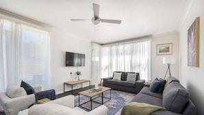 Ein modernes Wohnzimmer mit einem grauen Sofa, beigen Sesseln, einem Couchtisch und einem an der Wand montierten Fernseher.