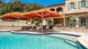 Ein Schwimmbecken mit Liegestühlen und Sonnenschirmen, umgeben von Steinmauern und einem Gebäude mit grünen Fensterläden.