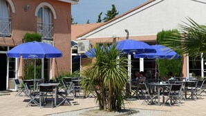 Ein Straßen-Café mit Tischen, Stühlen, blauen Sonnenschirmen und einem Gebäude mit einem Ziegelbedachung.