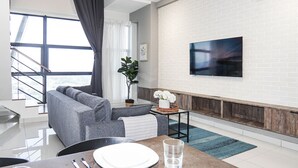 A modern living room with a grey sofa, a dining table with a glass, and a flat-screen TV mounted on a white brick wall. - Arte Mont Kiara Managed by Airhost (Kuala Lumpur)