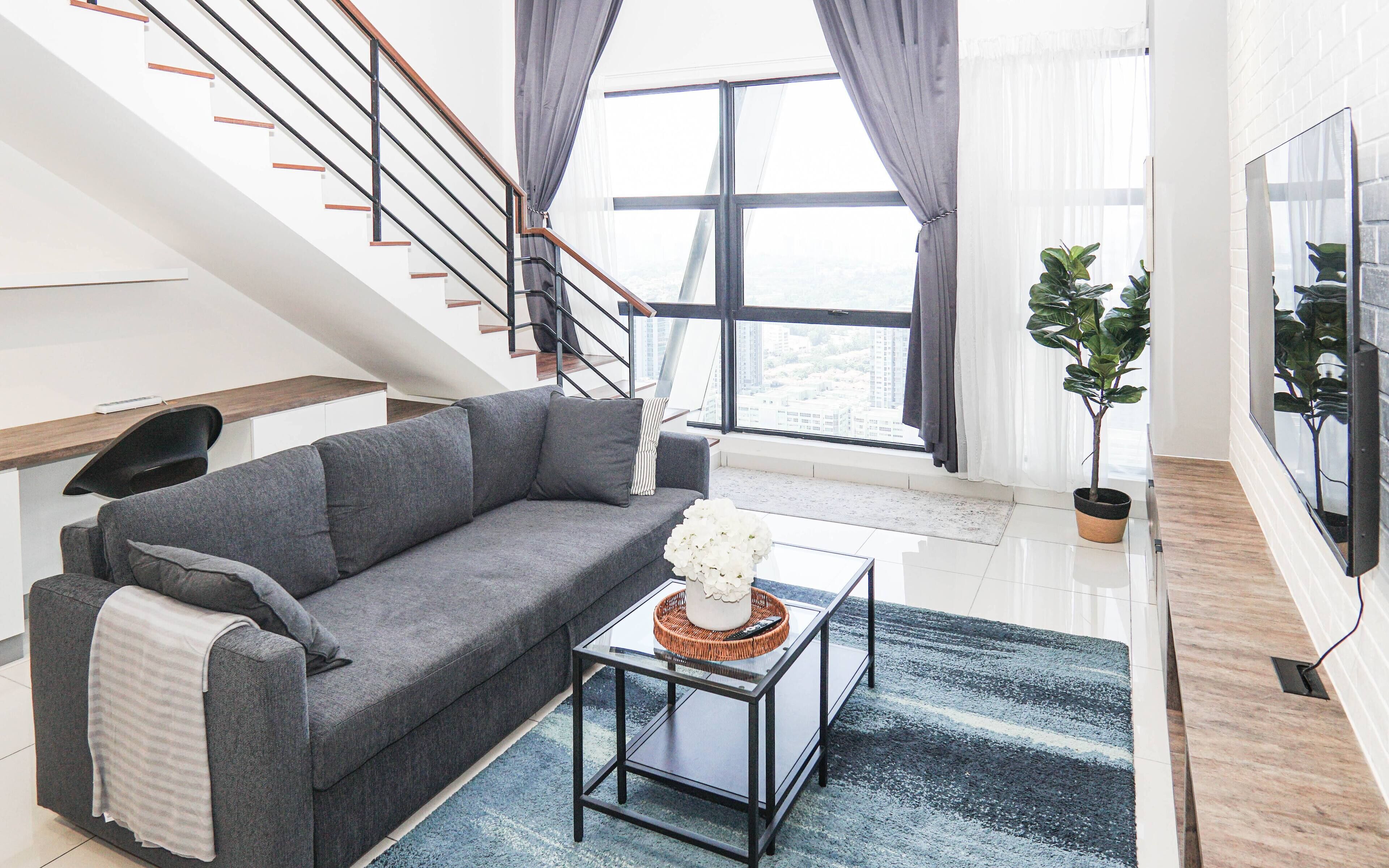 A modern living room with a grey sofa, a coffee table with a decorative item, a staircase with black railings, and a large window with curtains.