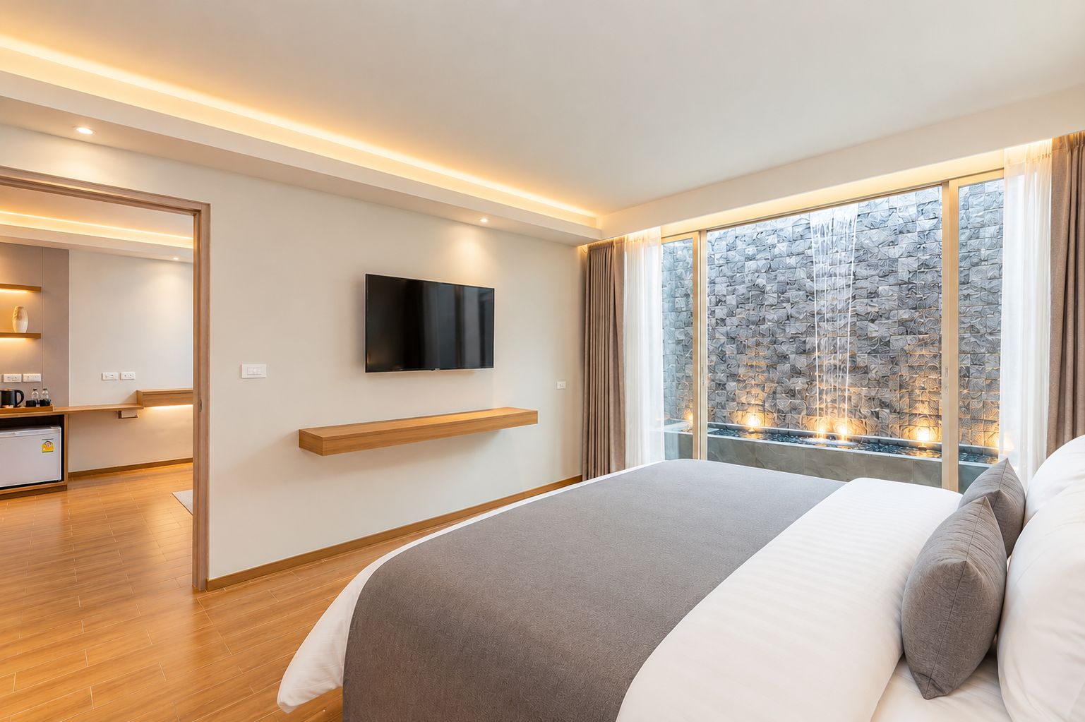 Una habitación de hotel moderna con una cama grande, un televisor de pantalla plana montado en la pared, un estante de madera y vistas a una pared de piedra por la ventana.