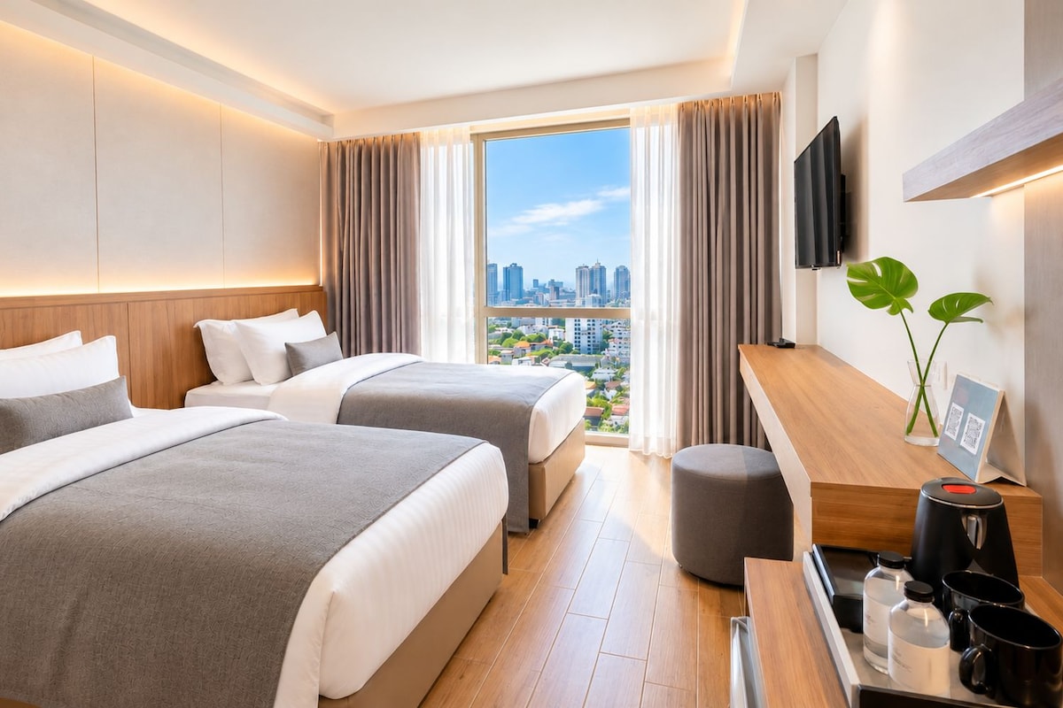 Habitación de hotel con dos camas, un escritorio con cafetera, televisor y vista a la ciudad.