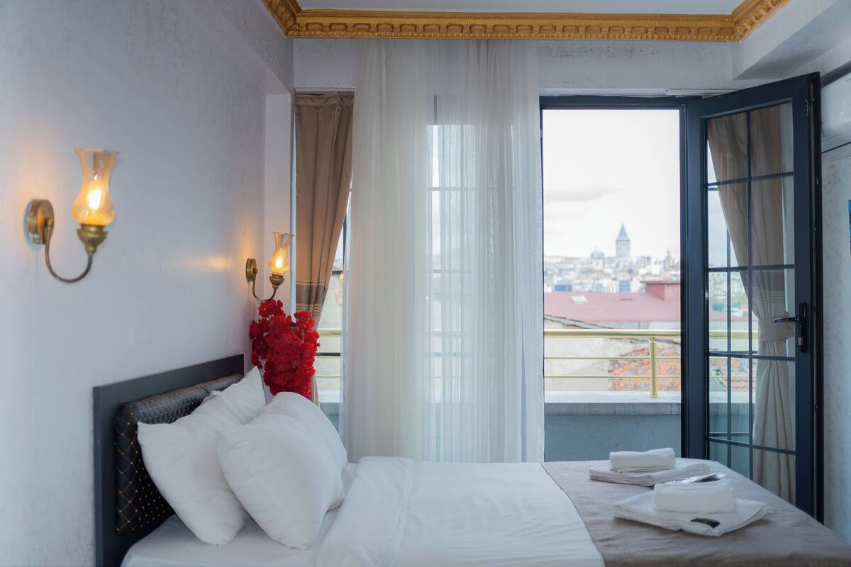 A hotel room with a bed, two wall sconces, a window with curtains, and a view of a cityscape.