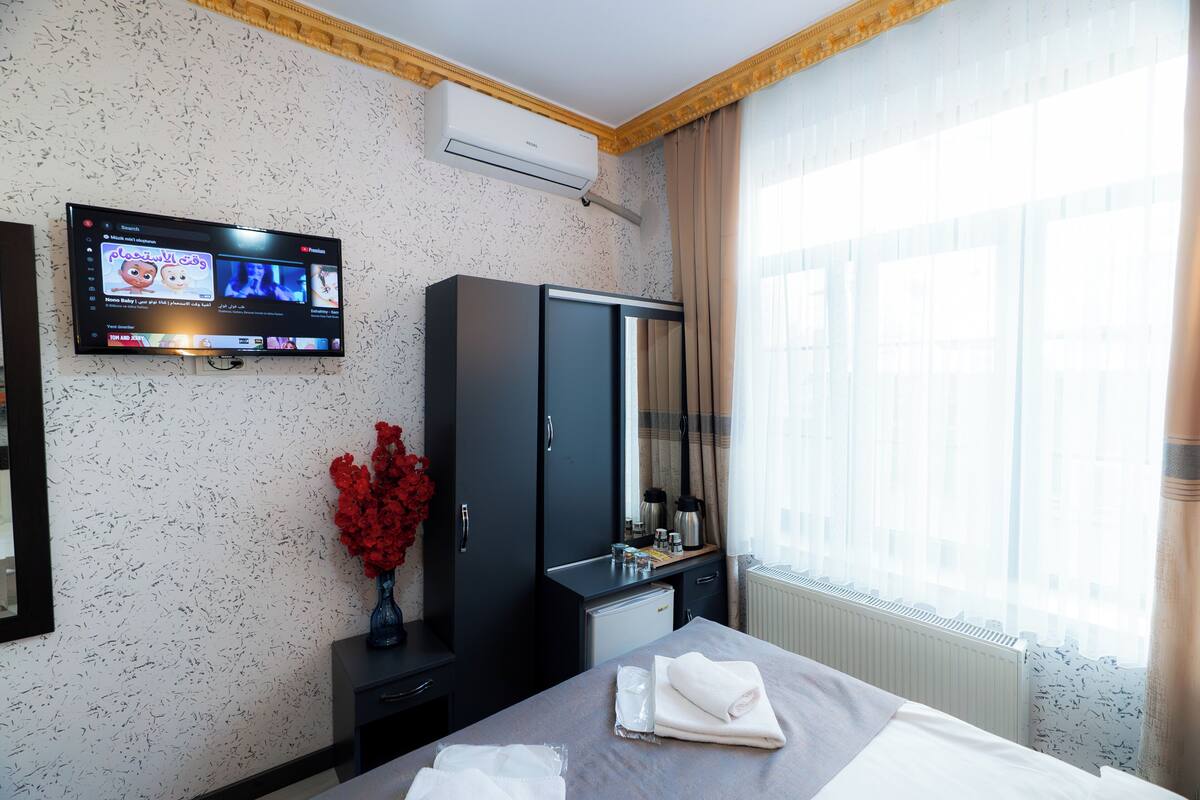 A hotel room with a bed, a TV mounted on the wall, a wardrobe, a small desk, a vase with flowers, and a window with curtains.