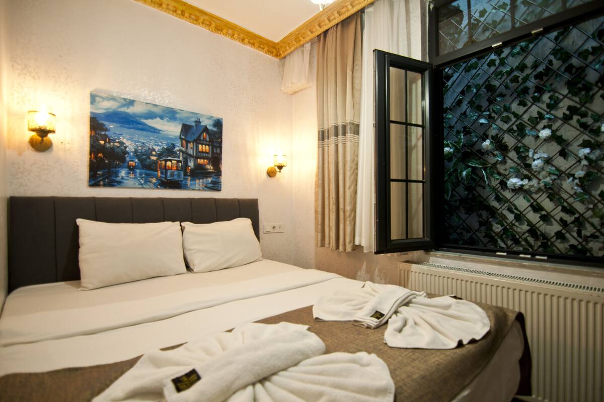A hotel room with a bed, two wall-mounted lights, a painting, a window with a view of greenery, and a radiator.