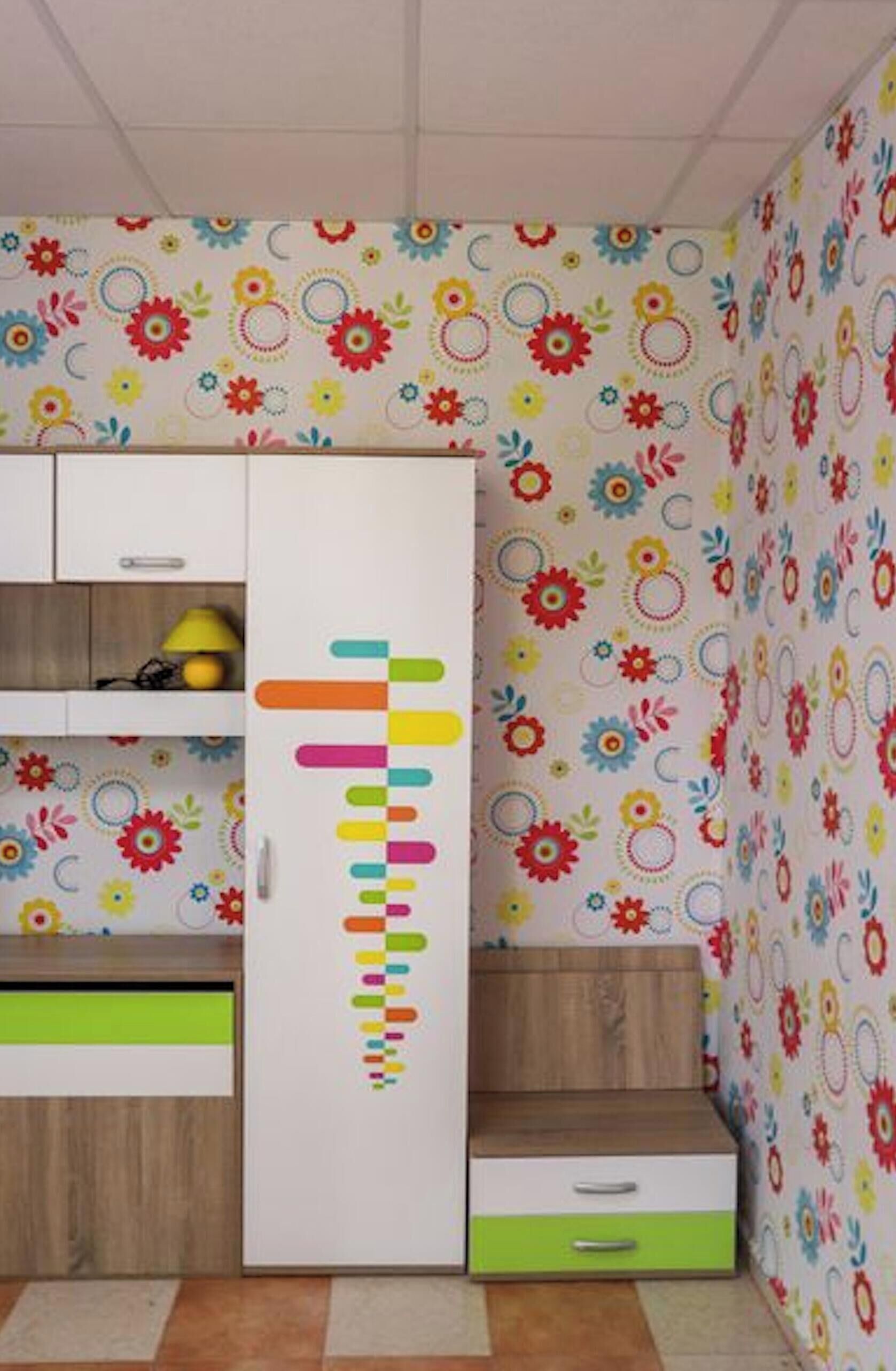 A room with a white wardrobe, a green drawer, a wooden bench, and a wall with a floral pattern.