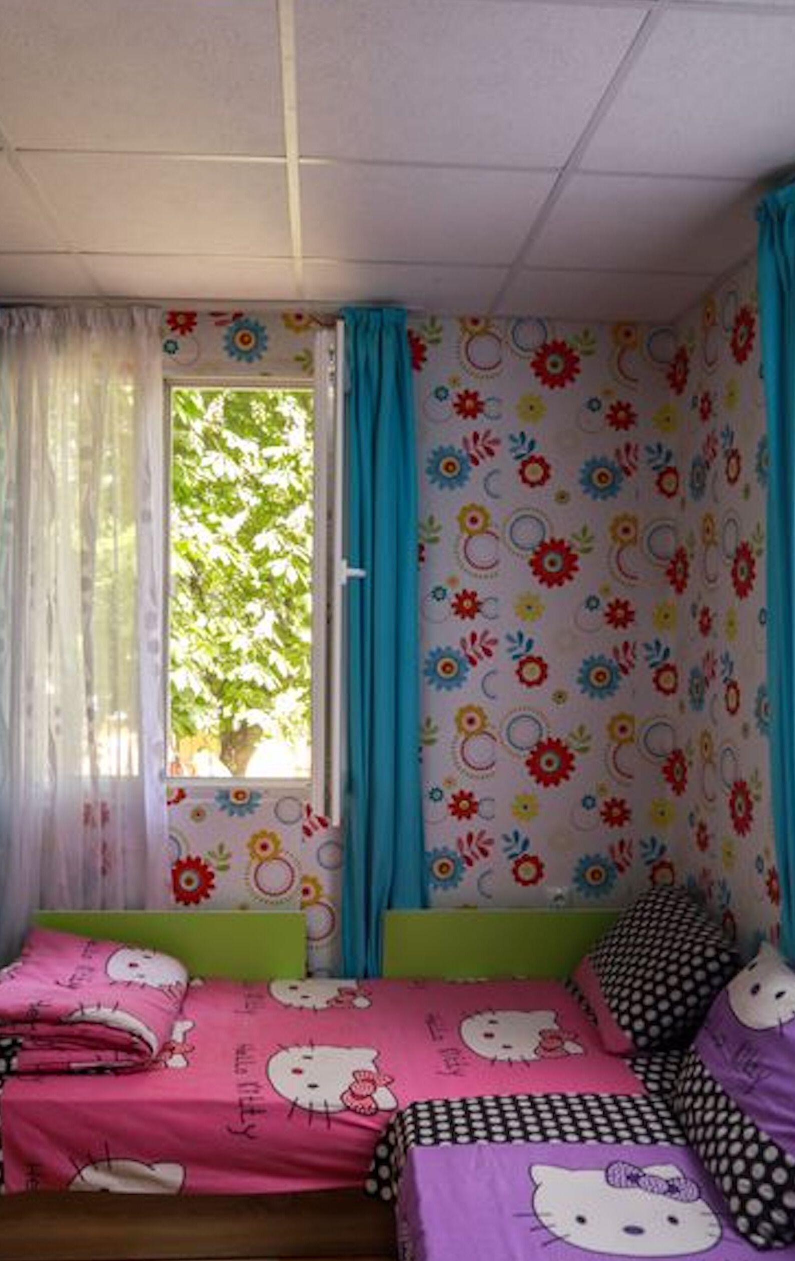 Ein Zimmer mit einem Bett, einem Fenster mit Vorhängen und einer Wand mit Blumenmuster.