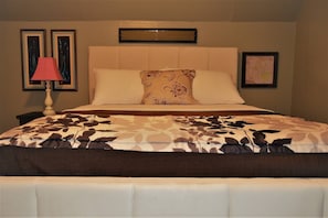 Uma cama bem arrumada com colcha estampada, um abajur e duas fotos emolduradas na parede.