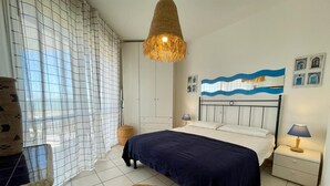 A bedroom with a bed, a hanging light fixture, a wall with a wave pattern, and a small table with a lamp. - Stunning Seafront Apartment (Caorle)