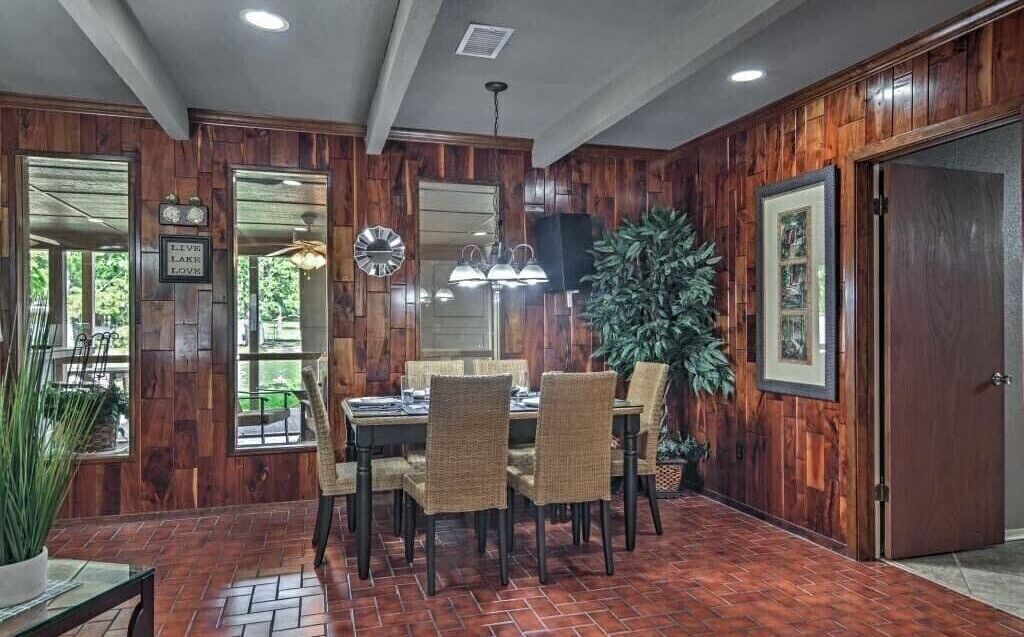 A dining area with a brick floor, wooden panel walls, a dining table with chairs, and a large potted plant.