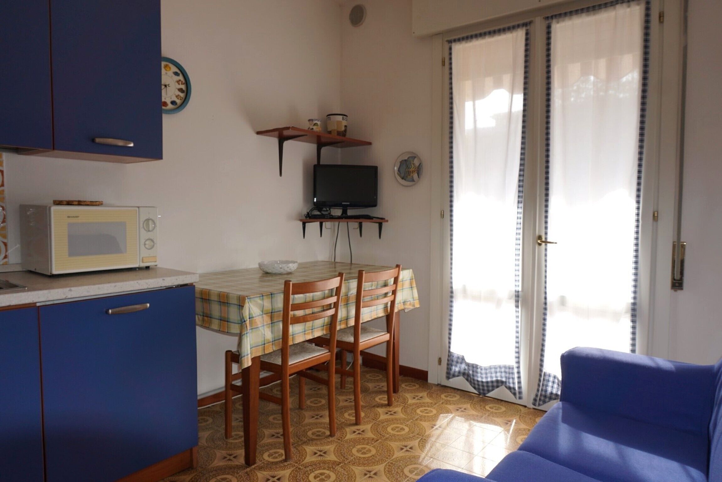 A compact kitchen with a microwave, a small TV on a stand, and a dining table with chairs. There is a blue sofa and a window with curtains.