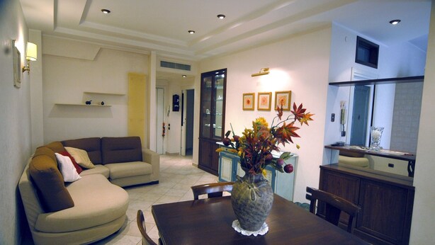 A living room with a sofa, a dining table with chairs, a vase with flowers, and a kitchen area with cabinets and a window.