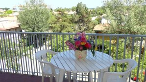 A balcony with a white table and chairs, a vase of flowers, and a view of greenery and buildings. - Cozy Flat With Balcony (Caorle)