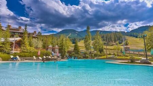 Ein klares, blaues Schwimmbecken mit Liegestühlen, umgeben von Bäumen und einer Bergkulisse.
