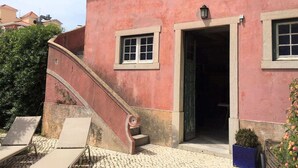 Ein pinkfarbenes Gebäude mit Treppe, einer Tür und einem Fenster. Bequeme Liegestühle und ein Topf mit einer Pflanze sind ebenfalls vorhanden.