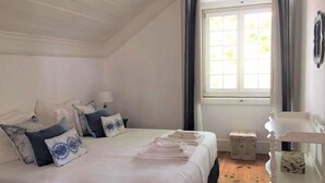 Ein Schlafzimmer mit einem großen Bett, einem Nachttisch, einem Fenster mit Vorhängen und einem kleinen Hocker.